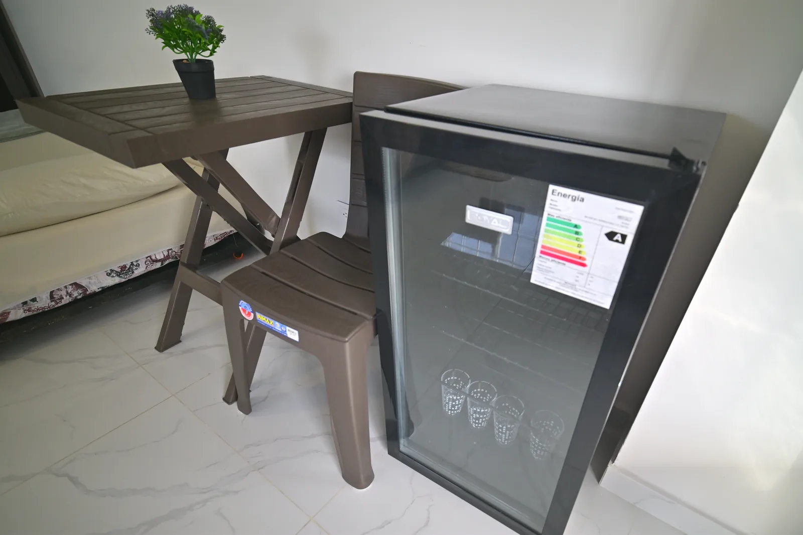 Minibar de puerta de vidrio con vasos adentro, junto a una mesa plegable y una silla.