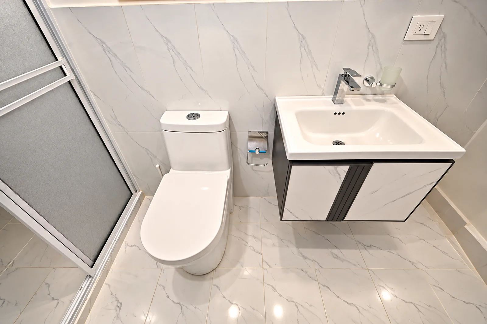 Baño de mármol blanco con lavamanos suspendido, sanitario y cabina de ducha de vidrio.
