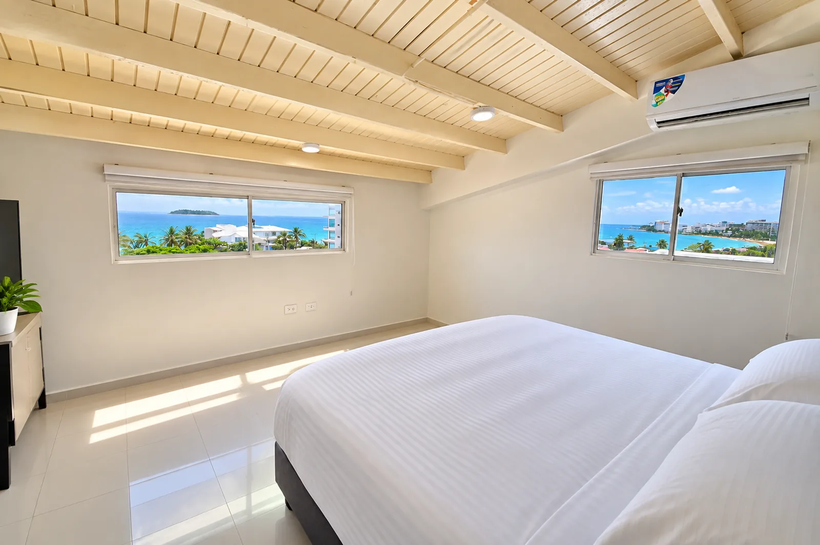 Habitación blanca de vigas de madera a la vista, con dos ventanas al mar a lado y lado de la cama.