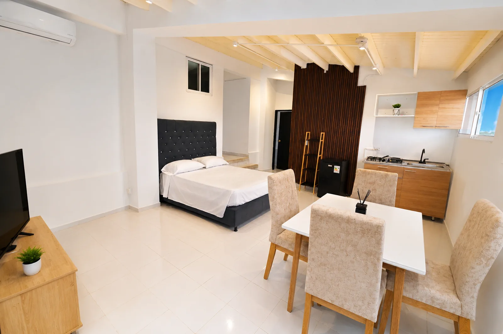 Apartamento con cama doble, comedor de cuatro puestos y cocina de madera separados por un panel de listones.