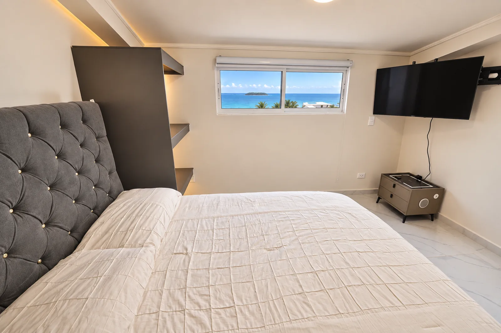 La cama de cabecero capitoneado junto a la columna de estantes, con el televisor y una ventana estrecha al mar.