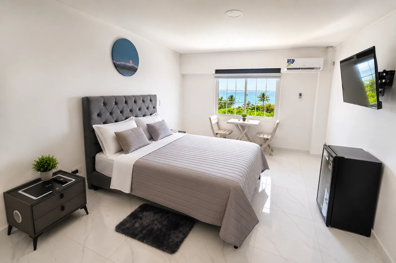 Habitación con cama doble de cabecero capitoneado, minibar y mesa para dos junto a la ventana, con palmeras y mar al fondo.