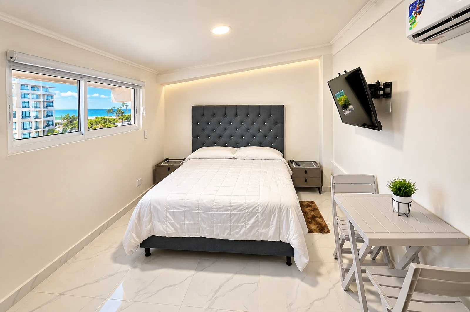 La misma habitación con la mesa de dos bajo la ventana, el televisor enfrente y el mar entre los edificios.