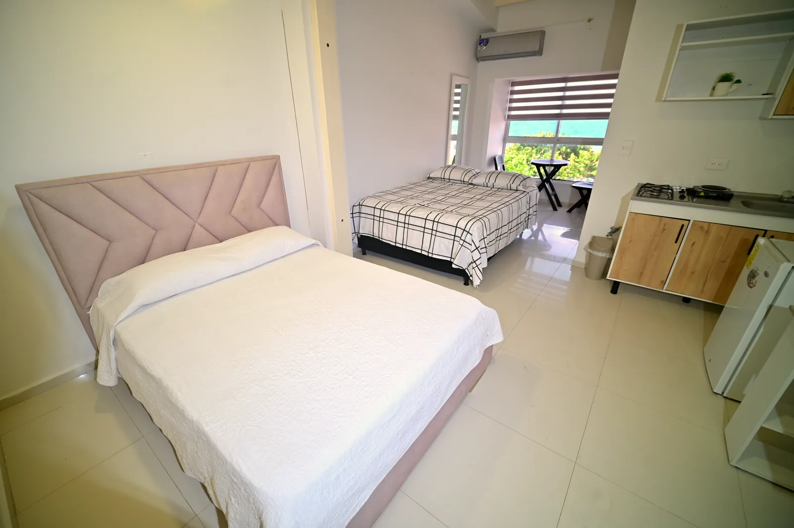 Estudio largo con una cama queen de cabecero rosado, una segunda cama al fondo, cocina con hornilla de gas y balcón al mar.